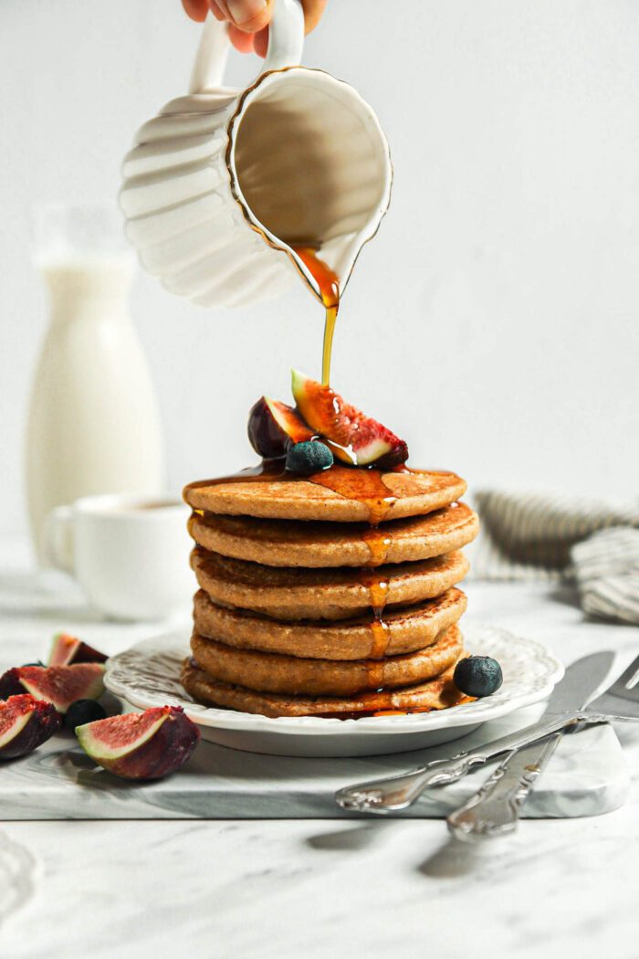 Hand pouring maple syrup from a small container over a stack of healthy oatmeal pancakes.