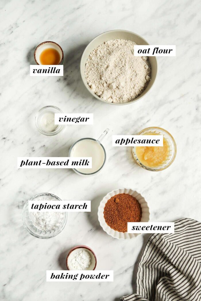 Oat flour, tapioca starch, sugar, vanilla and baking powder in small bowls on a marble surface. Each ingredient is labelled with text.