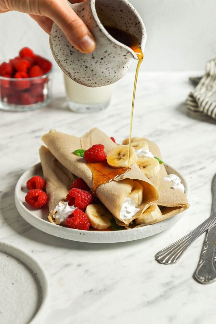 Hand pouring maple syrup from a small container over a stack of buckwheat crepes filling with strawberries and banana on a plate.