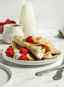 3 buckwheat crepes filling with sliced strawberries and banana and topped with maple syrup on a plate.