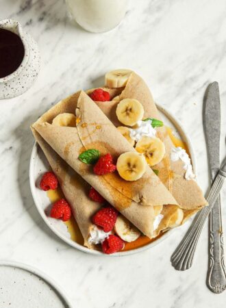 Overhead view of 3 buckwheat crepes filled with strawberries, banana and whipped cream on a plate.