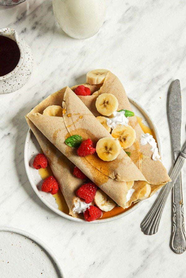 Overhead view of 3 buckwheat crepes filled with strawberries, banana and whipped cream on a plate.