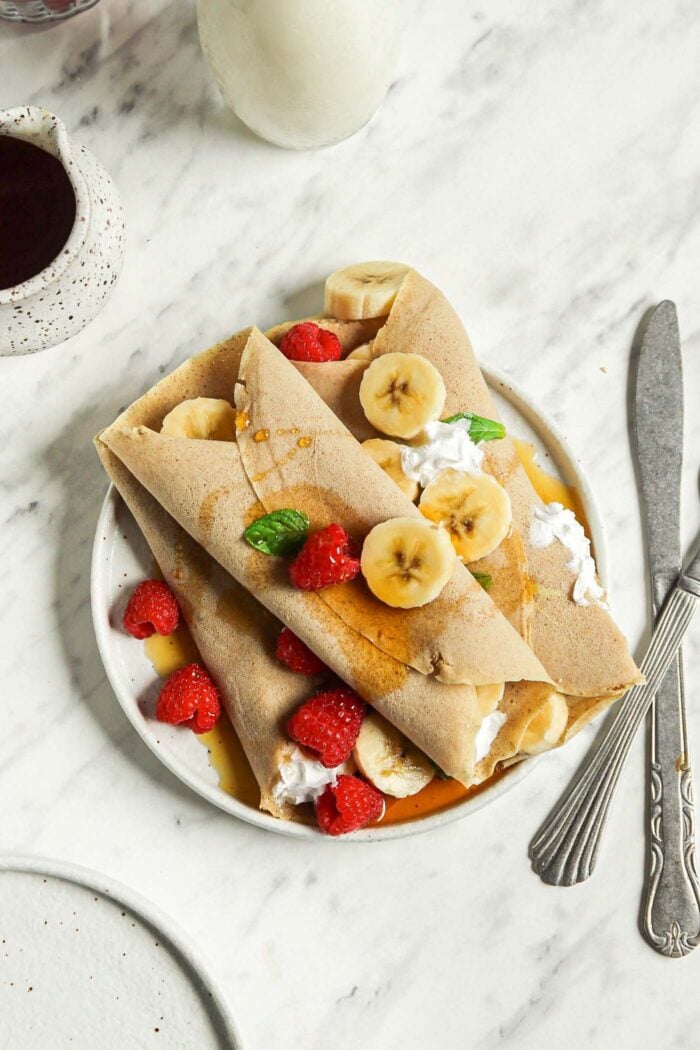 Overhead view of 3 buckwheat crepes filled with strawberries, banana and whipped cream on a plate.