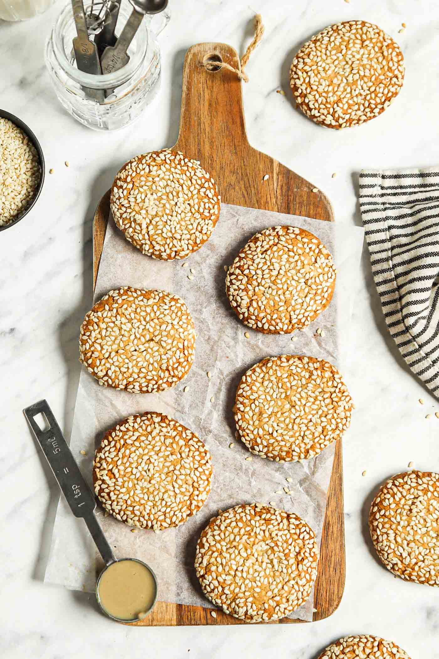 A number of sesame cookies on a wooden cutting board with with a measuring spoon of tahini resting on the edge of the board and a few more cookies scattered around it.