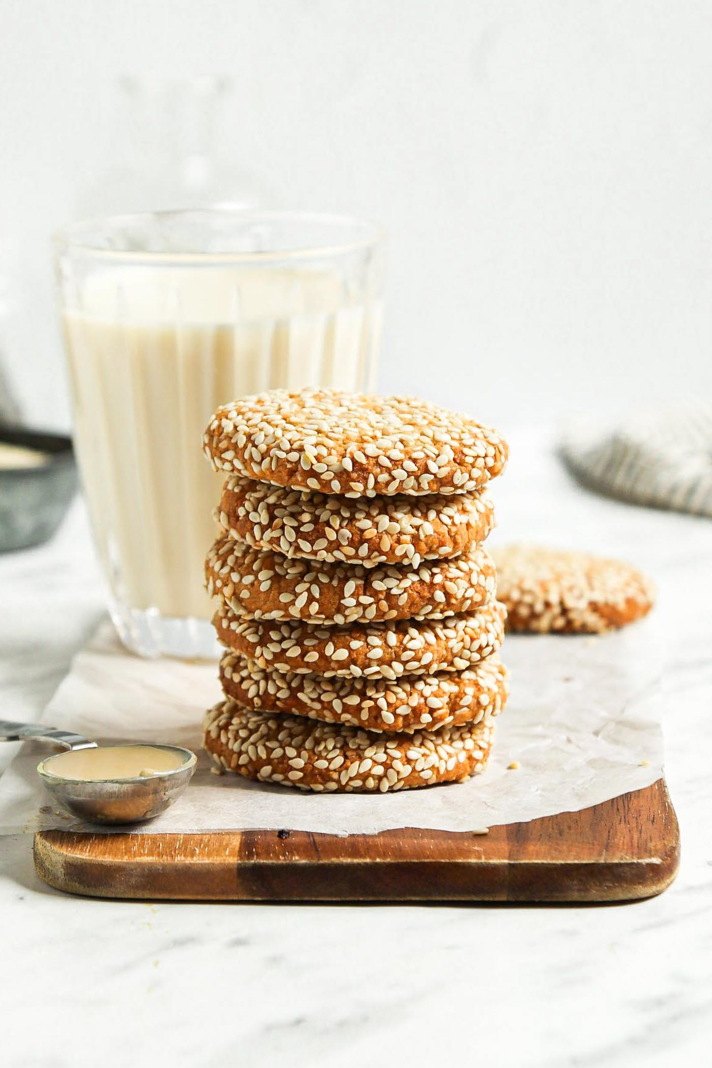 Stack of 5 sesame seed-coated cookies on a cutting board with a glass of milk behind them.