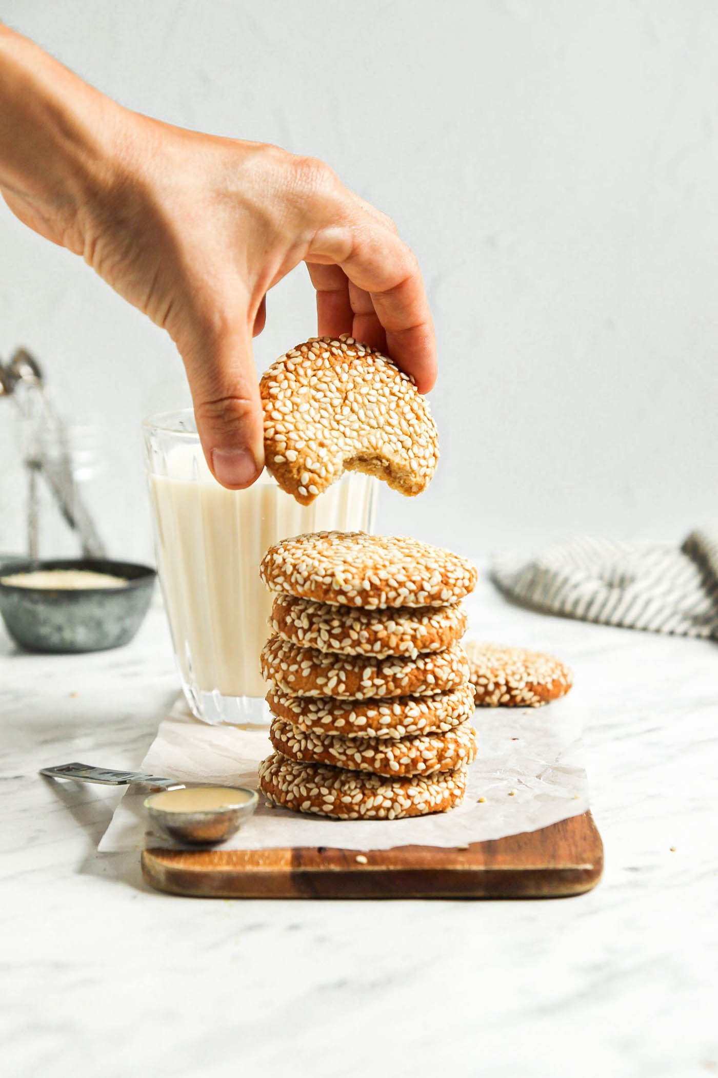 Hand lifting a sesame seed-coated tahini cookie from a stack of cookies on a cutting board.