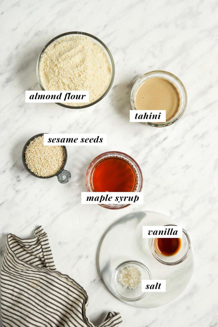 Almond flour, tahini, maple syrup, sesame seeds, vanilla and salt in small bowls on a marble surface. Each ingredient is labelled with text.