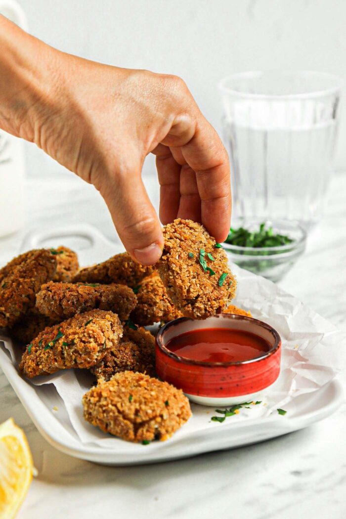 Hand dipping a baked tofu nugget in a small bowl of ketchup.