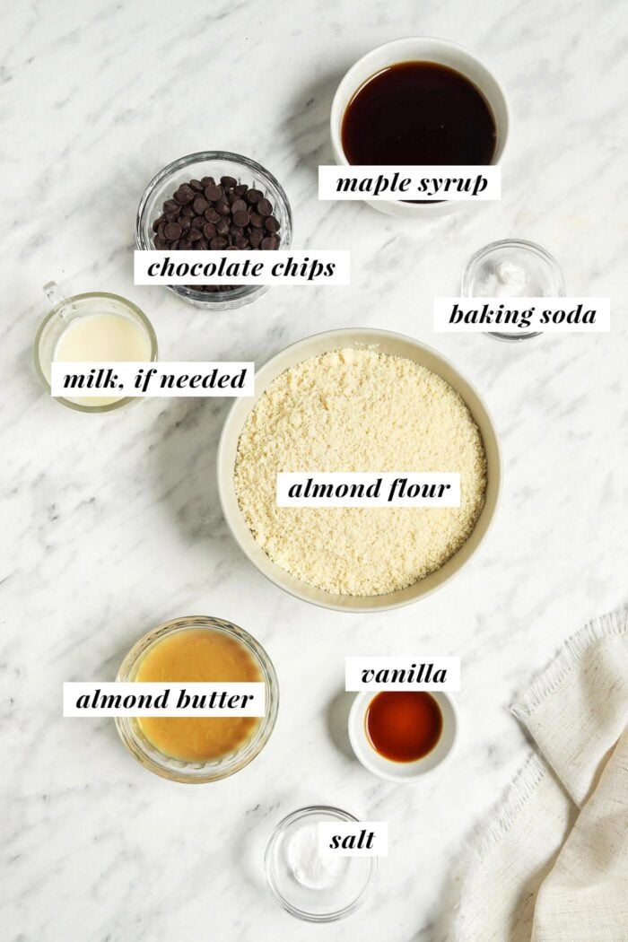 Todos los ingredientes necesarios para hacer una receta de galleta con chispas de chocolate con harina de almendras en tazones pequeños sobre una superficie de mármol.