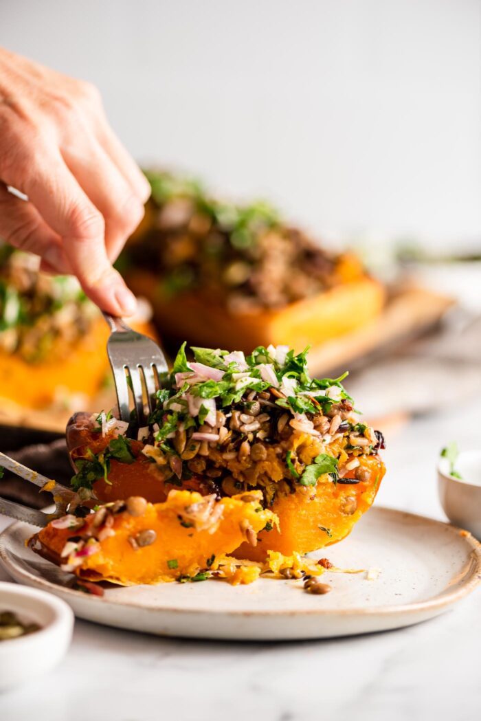 Hands using a knife and fork to slice a stuffed butternut squash on a plate.