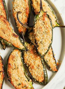 Plate of vegan baked jalapeno poppers sitting on cutting board beside small bowls of breadcrumbs and hot sauce.
