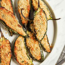 Plate of vegan baked jalapeno poppers sitting on cutting board beside small bowls of breadcrumbs and hot sauce.