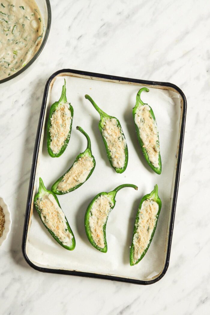 Jalapenos filled with a cashew cream and topped with breadcrumbs on a baking tray lined with parchment paper.