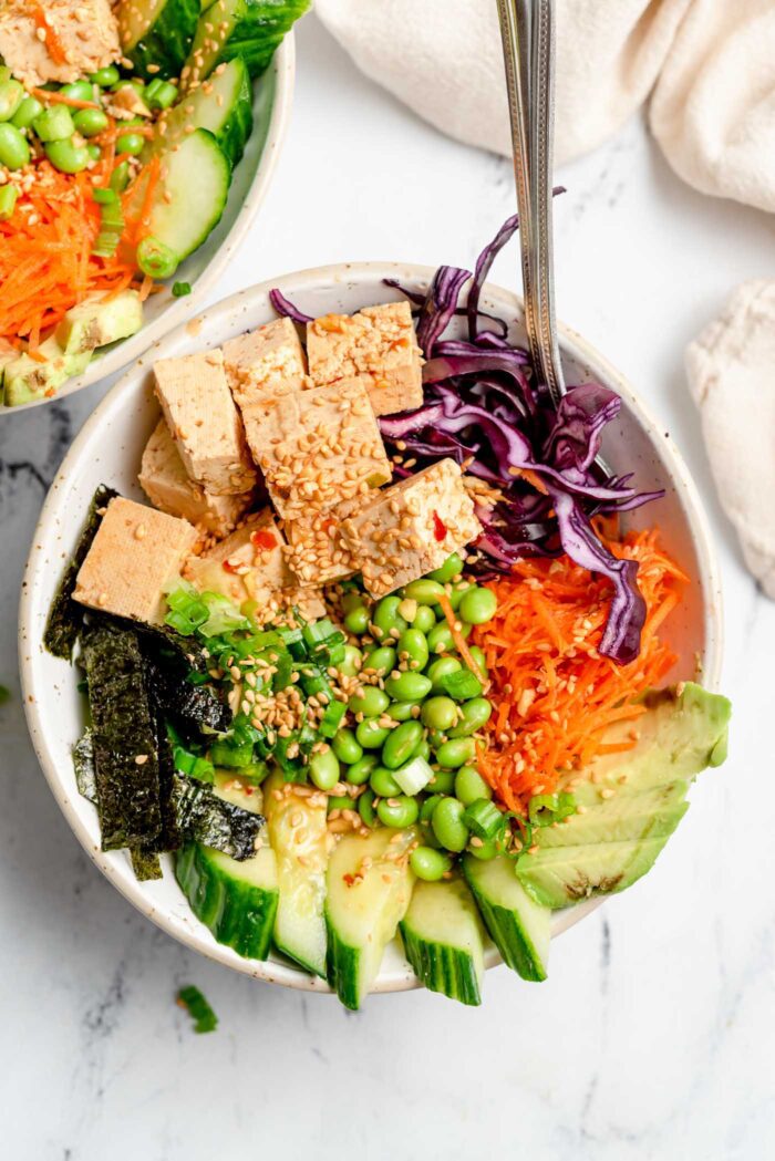Vista superior de un tazón de poke de tofu vegano con zanahoria, aguacate, zanahoria, repollo, edamame, sésamo y pepino.