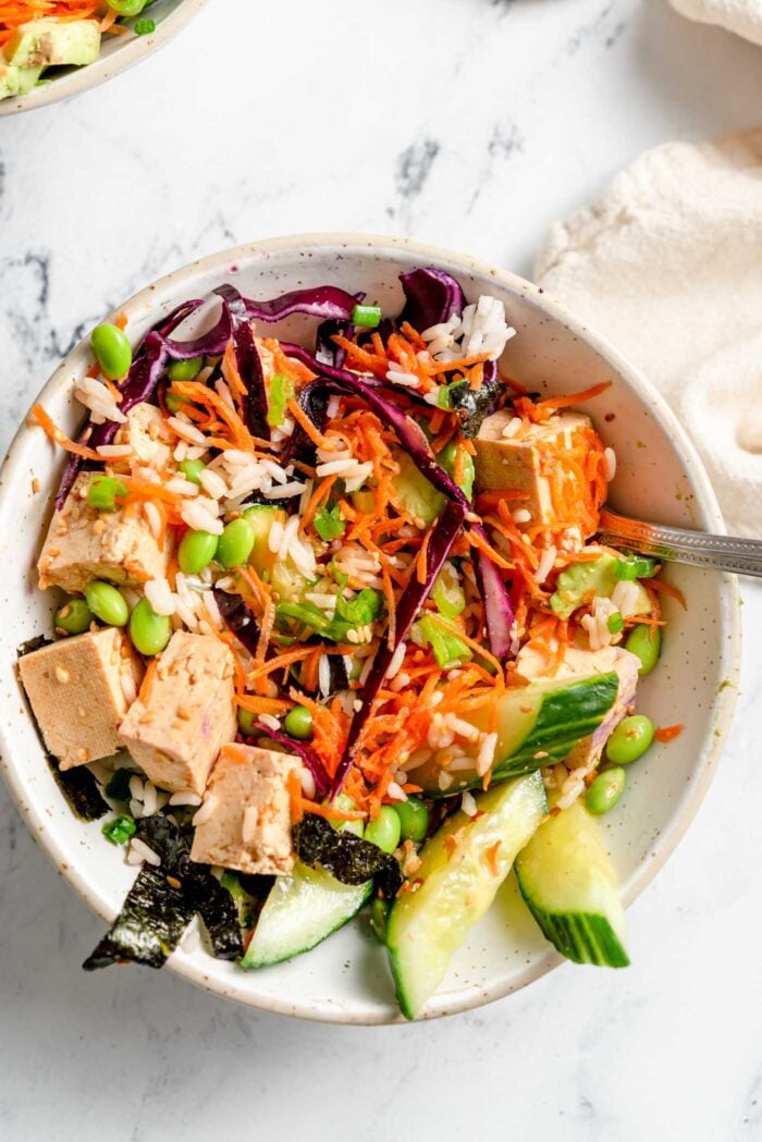Rice, tofu, cucumber, edamame, cabbage, carrot, sesame seeds and nori strips mixed up in a bowl with a fork in it.