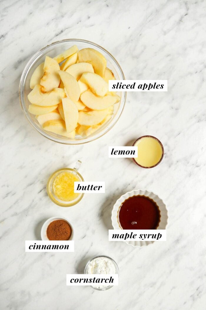 Apples, lemon, butter, maple syrup, cinnamon and cornstarch in bowls on a marble counter.