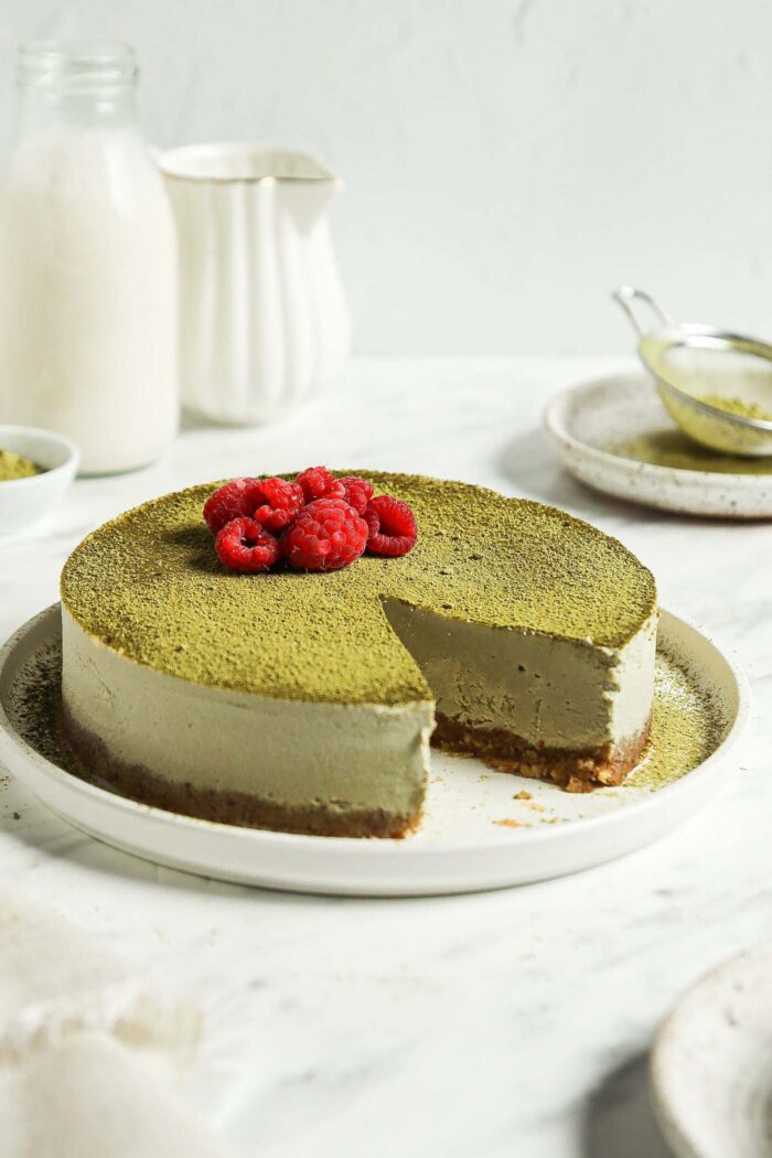 Raw no-bake matcha cheesecake topped with matcha powder and raspberries on a plate.
