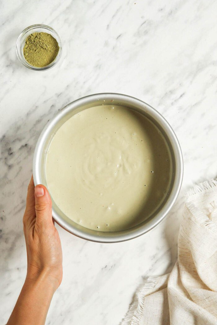 Raw no-bake matcha cheesecake in a round springform. A hand is holding the edge of the pan.