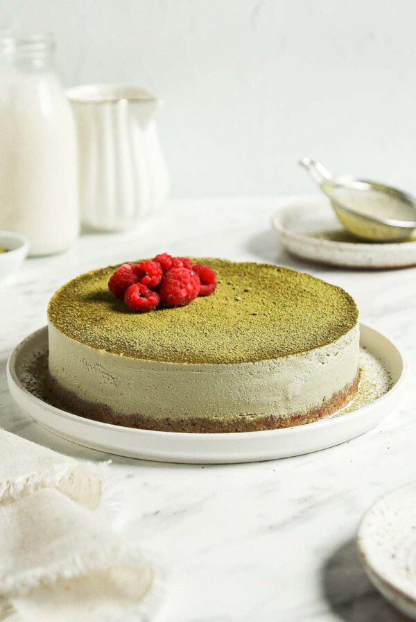 Raw no-bake matcha cheesecake topped with matcha powder and raspberries on a plate.