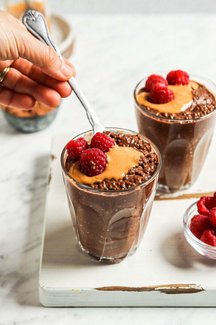 Two bowls of chocolate chia seed pudding topped with peanut butter and fresh raspberries. Hand is using a spoon in one of the bowls.