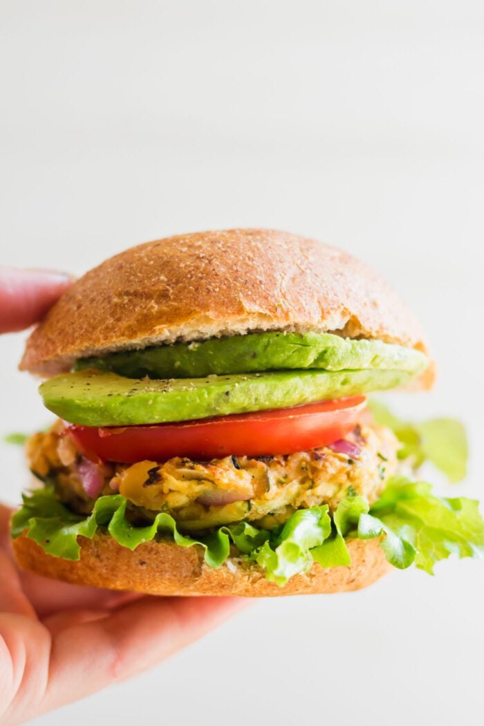 Hand holding a chickpea veggie burger on a bun with lettuce, tomato, onion and avocado.