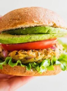 Hand holding a chickpea veggie burger on a bun with lettuce, tomato, onion and avocado.