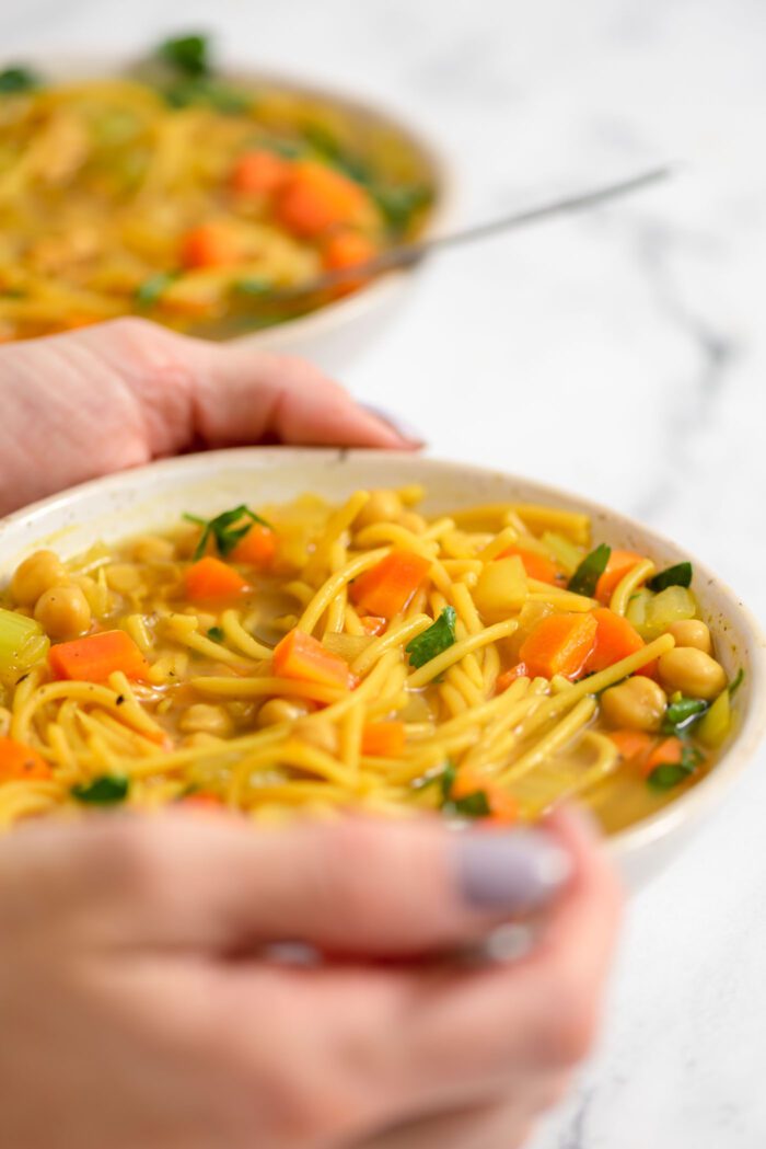 Two hands holding a bowl of vegan chickpea noodle soup with carrot and celery.