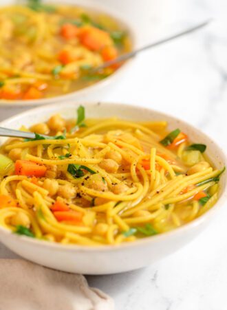 Two bowls of vegan chicken noodle soup with vegetables and spaghetti noodles.