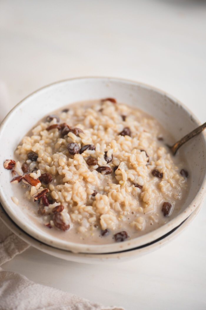 Bowl of brown rice pudding with raisins and pecans. A spoon rests in the bowl.