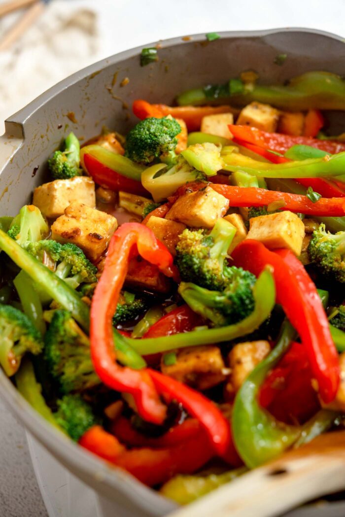 Cubed tofu and stir fried vegetables cooking in a stick orange sauce in a pan.