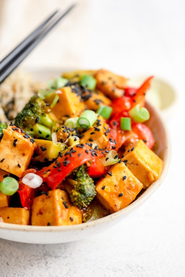 Pair of chopsticks rests on a bowl of stir fried vegetables and tofu in orange sauce.