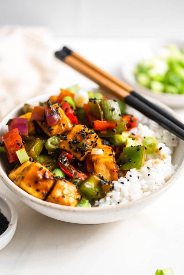 A pair of chopsticks rests on a bowl of sweet and sour stir fry with vegetables served over rice and topped with sesame seeds and green onion.