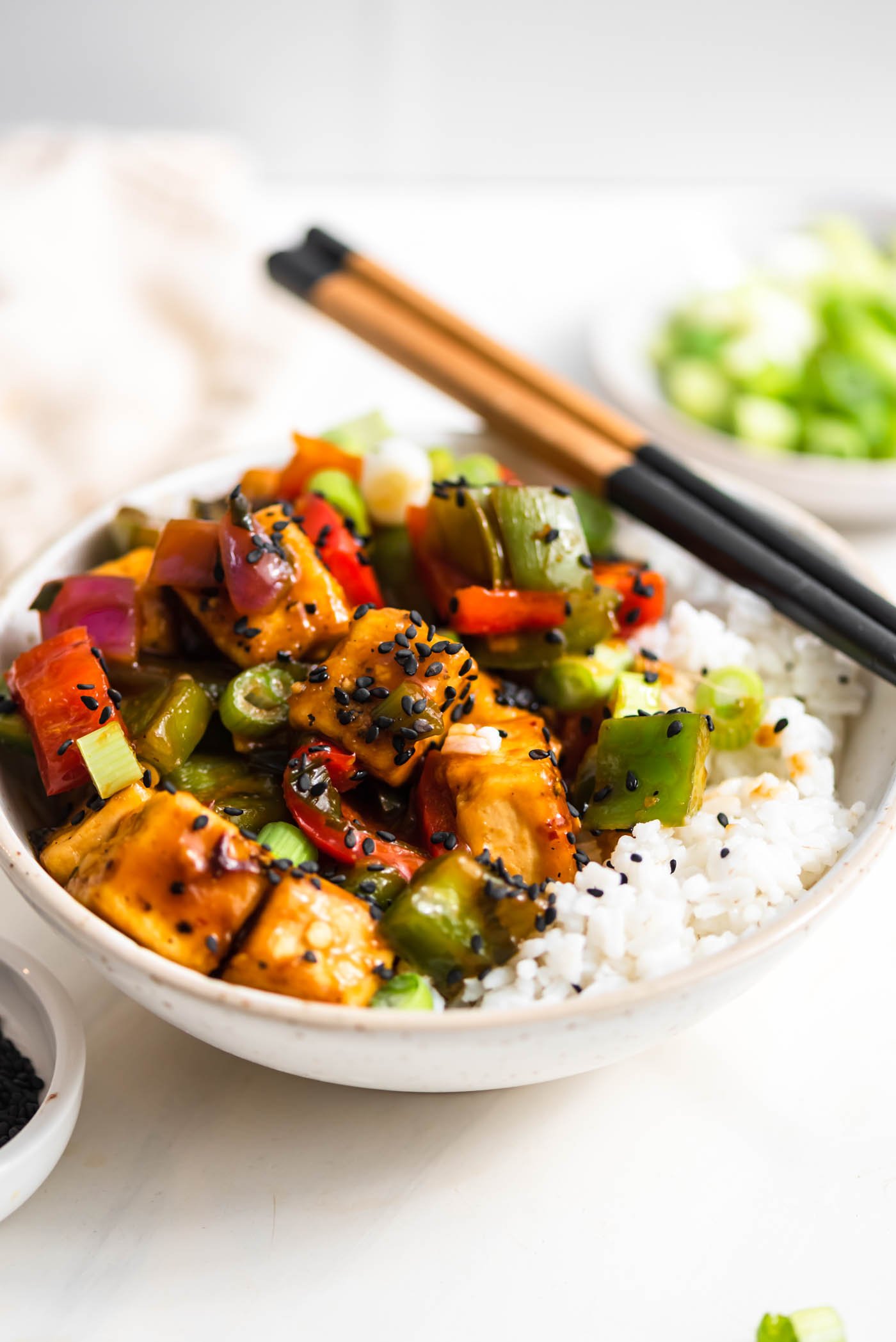 A pair of chopsticks rests on a bowl of sweet and sour stir fry with vegetables served over rice and topped with sesame seeds and green onion.