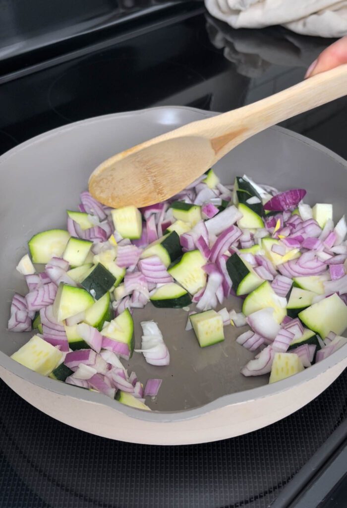 Chopped onion and zucchini cooking in a skillet with a wooden spoon.