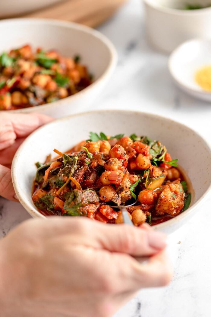 Hand using a spoon in a bowl of Mediterranean tomato chickpea stew with spinach.