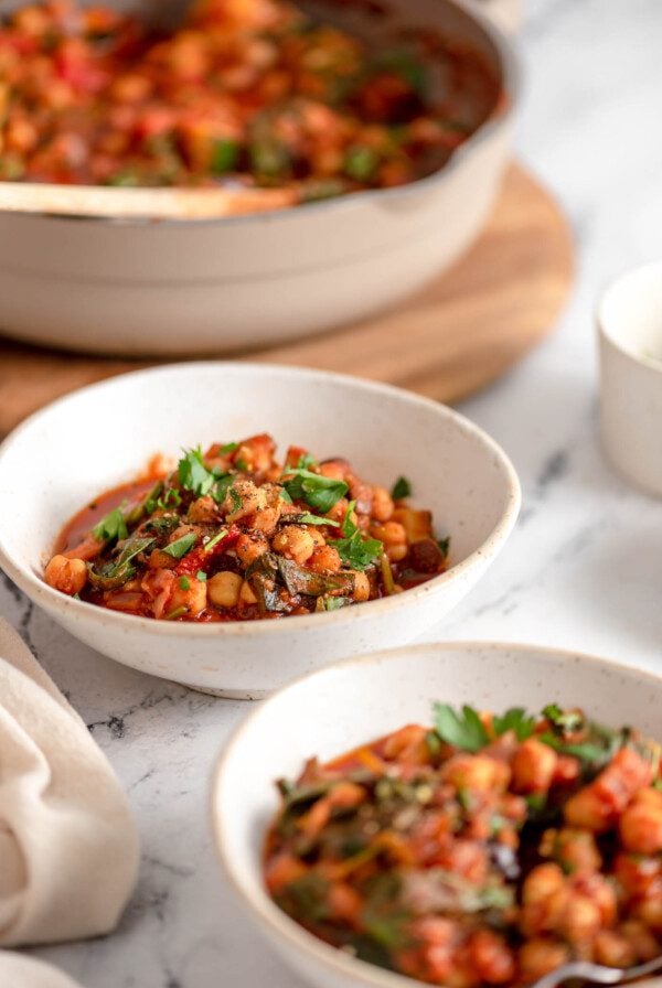 Two bowls of tomato chickpea stew with spinach. A skillet of leftover stew is show in the background.