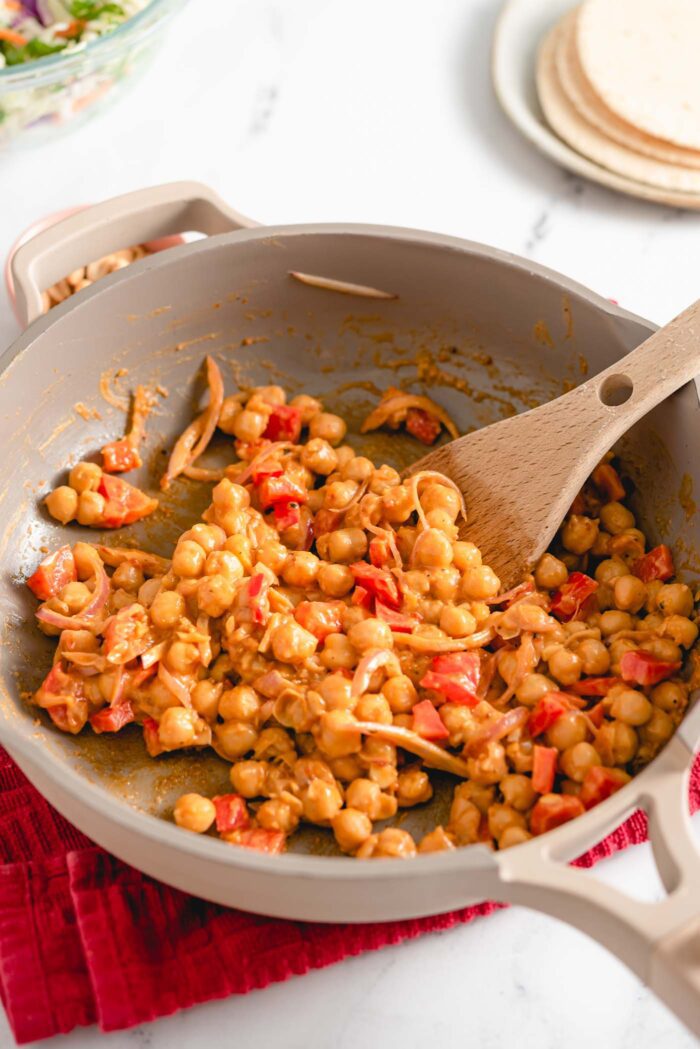 Chickpeas, red onion and shallots cooking in peanut sauce in a skillet with a wooden spoon.