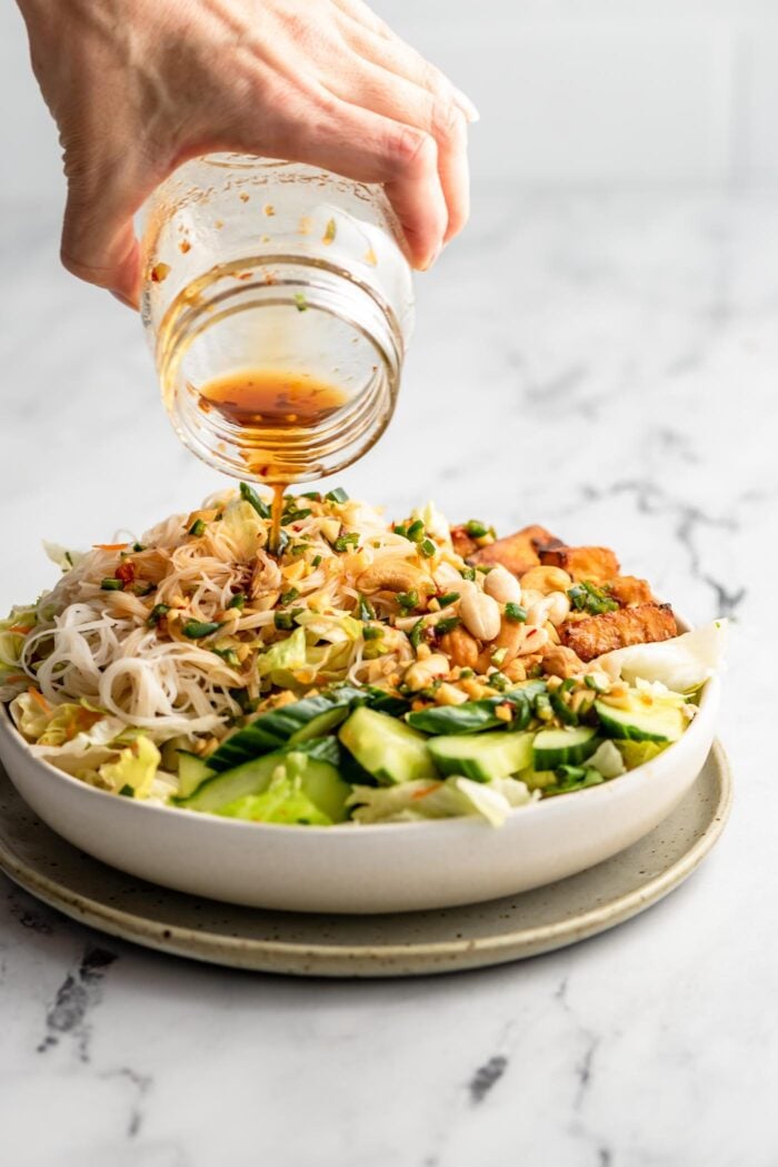 Pouring salad dressing over a vermicelli noodle salad with peanuts and cucumber.