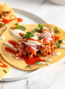 Three vegan tempeh tacos topped with pickled onion, cashew crema and hot sauce on a plate.