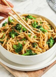 Bowl of sesame noodles with broccoli topped with scallions and sesame seeds. A pair of chopsticks rests on the bowl.