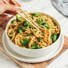 Bowl of sesame noodles with broccoli topped with scallions and sesame seeds. A pair of chopsticks rests on the bowl.