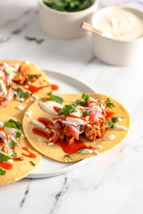 Three vegan tempeh tacos topped with pickled onion, cashew crema and hot sauce on a plate.