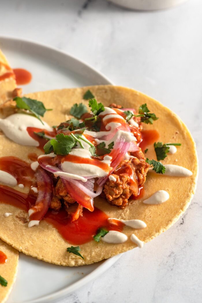 Close up of a tempeh taco topped with pickled red onion, cilantro, cashew cream sauce and hot sauce.