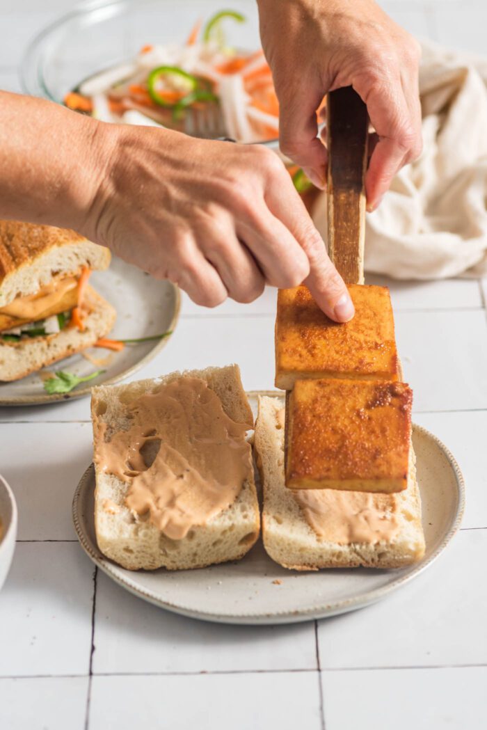 Adding thick slices of baked tofu to a toasted baguette slathered in spicy mayo sauce.