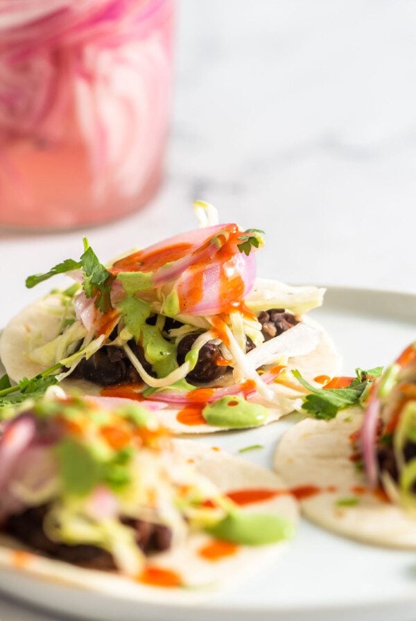 A small soft taco topped with pickled red onion, cabbage and cilantro crema on a plate. Two more tacos are blurred in the foreground.