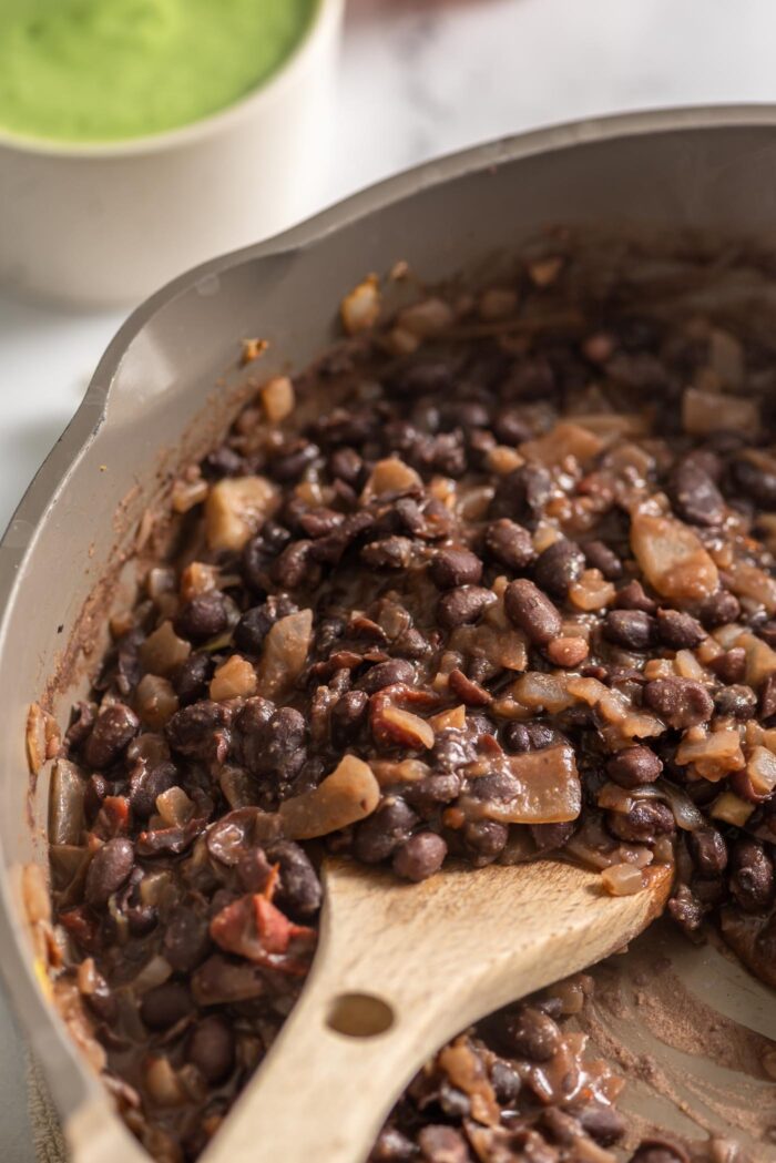 Cooked black beans and onions in a skillet with a wooden spoon.