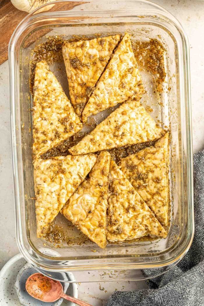 Thick slices of tempeh being marinated in a herb balsamic seasoning in a glass food storage container.