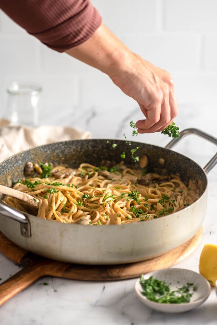 Hand sprinkling chopped parsley over a large skillet of vegan carbonara with linguini noodles and mushrooms.