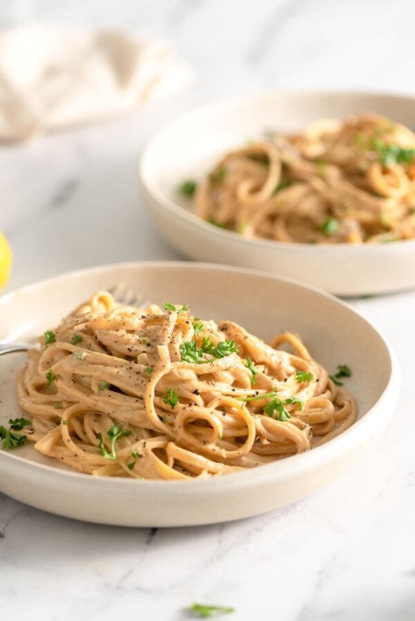 Two bowls of vegan carbonara pasta topped with pepper and chopped parsley in shallow bowls on a marble surface.