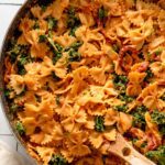 Creamy sun-dried tomato bowtie pasta with kale in a large skillet with a wooden spoon.